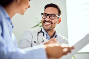 A smiling male doctor wearing glasses and a stethoscope talks with a patient, holding a clipboard in a bright, well-lit office, discussing pain management options that go beyond medication.