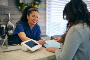 A smiling woman in medical scrubs hands a clipboard and pen to another woman at a reception desk, welcoming patients for pain management, with a payment terminal and floral wreath in the background.