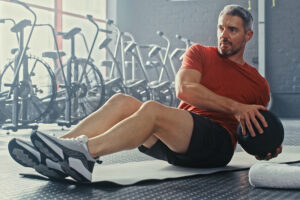 A man in a red shirt and black shorts does Russian twists with a medicine ball while sitting on a mat in a gym, focusing on functional fitness, with exercise bikes in the background.