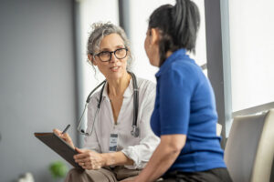 A female doctor with a stethoscope and clipboard smiles and talks with a female patient in a brightly lit medical office, both seated and engaged in conversation about Whole Person Care.