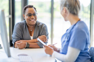 A woman smiles while sitting at a desk across from a healthcare professional in scrubs, who is holding a tablet and pen. They appear to be discussing the root cause of her recurring pain in a friendly, bright office setting.