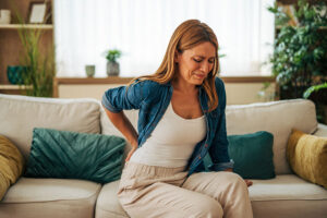 A woman sitting on a couch holds her lower back, appearing to experience recurring pain. She is wearing a denim shirt and light-colored pants, with home decor and plants in the background.