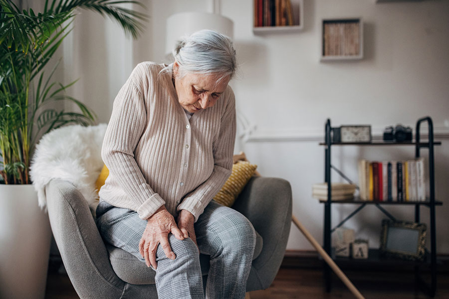An elderly woman with gray hair sits in a chair, holding her knee and appearing to be in pain, perhaps seeking pain treatment for recurring pain. She is indoors in a cozy living room with bookshelves, plants, and soft lighting.