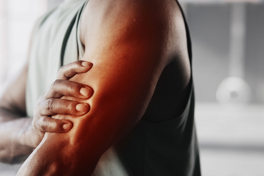 A person holds their upper arm with one hand, indicating recurring pain or discomfort. The arm is highlighted with a reddish tint, suggesting inflammation or injury. The background is blurred.