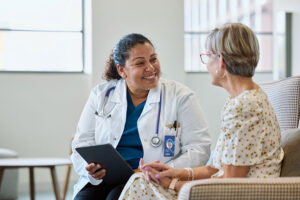 A doctor with a stethoscope smiles while discussing pain symptoms with a seated older woman in a light-patterned dress. The doctor holds a tablet, and they appear to be having a friendly, reassuring conversation in a bright room.