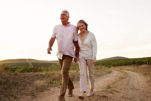 An older couple walks hand in hand along a dirt path through a field at sunset, smiling and enjoying each other's company despite occasional leg numbness, appreciating life while exploring causes and treatment options together.