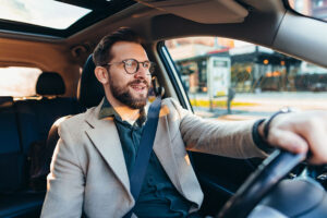 A man wearing glasses and a beige coat smiles while driving a car, enjoying his auto injury recovery after a recent car accident. Sunlight streams through the window, and the background shows an urban street scene.