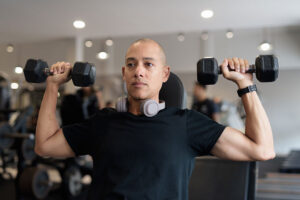 A person wearing a black shirt and headphones around their neck lifts two dumbbells while seated in a gym, focusing on their workout and taking care to avoid shoulder pain.