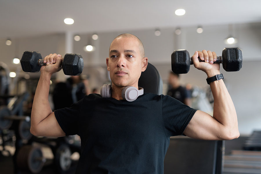 A person wearing a black shirt and headphones around their neck lifts two dumbbells while seated in a gym, focusing on their workout and taking care to avoid shoulder pain.