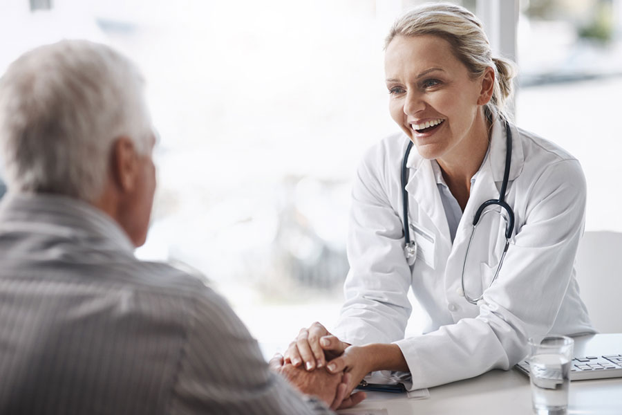 A smiling doctor with a stethoscope talks to an older male patient, gently holding his hand in a bright medical office, as they discuss herniated disc symptoms and ways to manage nerve compression.