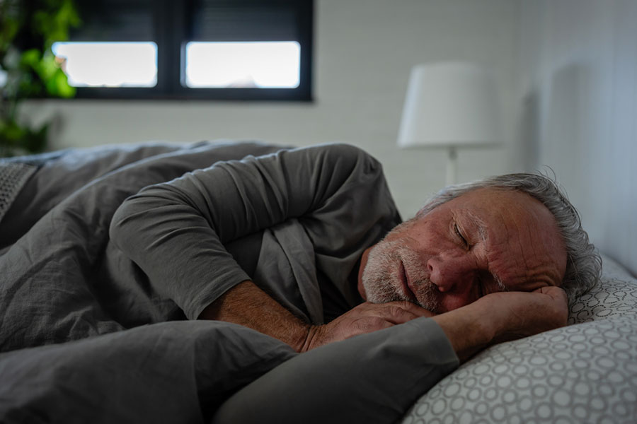 An older man with gray hair and a beard sleeps peacefully on his side in bed, covered with a gray blanket. Despite the softly lit lamp and a window in the background, he appears undisturbed by any nighttime pain—a contrast to those whose pain gets worse at night and are searching for reasons for pain at night.