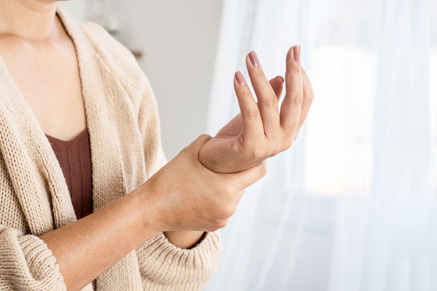 A person wearing a beige sweater holds their right wrist with their left hand, suggesting discomfort or pain—possibly due to hand numbness or the sensation of their hand falling asleep—in a softly lit room.