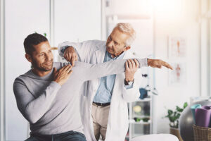 A doctor in a white coat examines a man’s shoulder and arm, gently extending the man’s arm while both are smiling in a well-lit medical office, discussing the symptoms location and whether there is any pain that travels or referred pain.