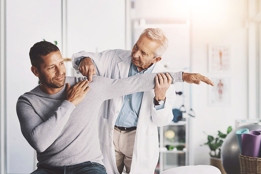 A doctor in a white coat examines a man’s shoulder and arm, gently extending the man’s arm while both are smiling in a well-lit medical office, discussing the symptoms location and whether there is any pain that travels or referred pain.